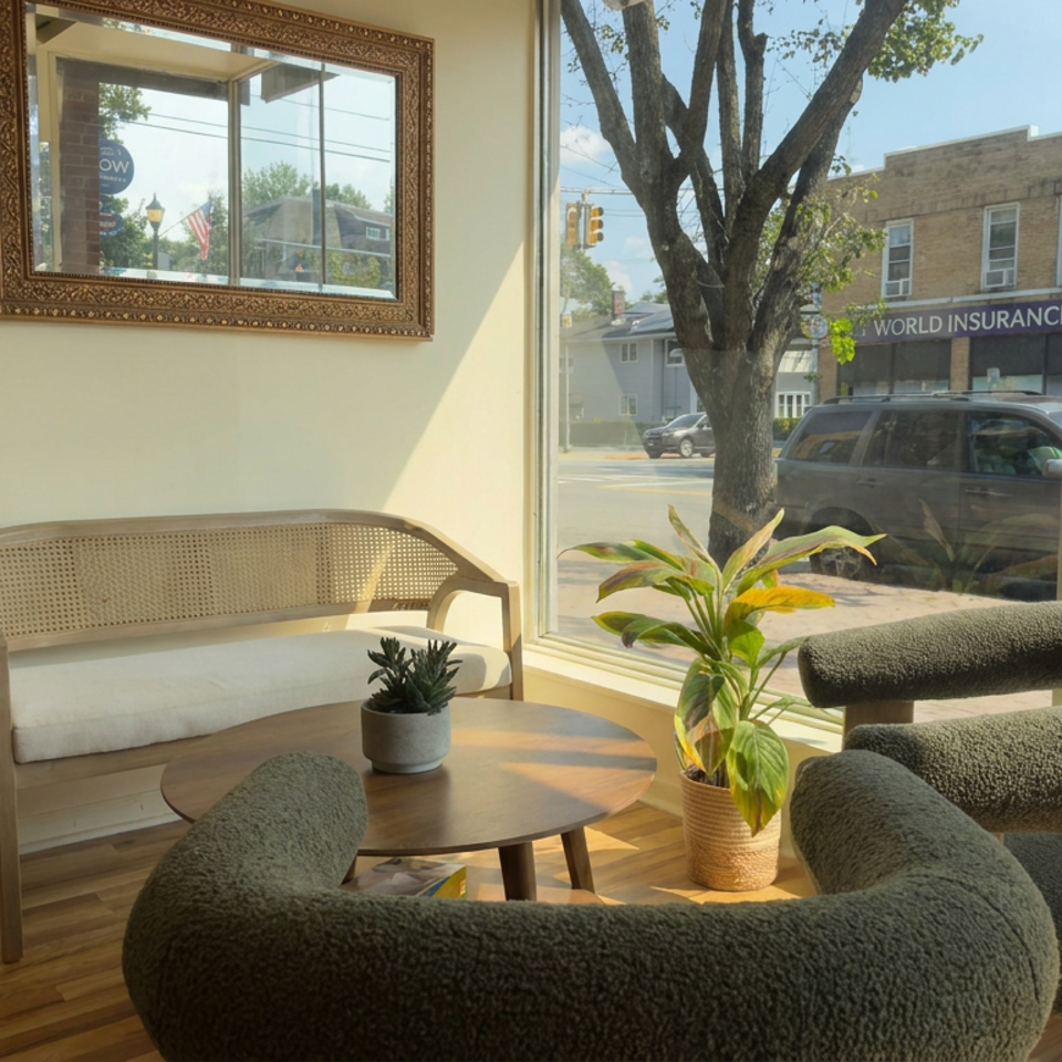 Sunlit restaurant seating area with curved upholstered chairs, a wooden table, bench, plants, and street views through large windows.
