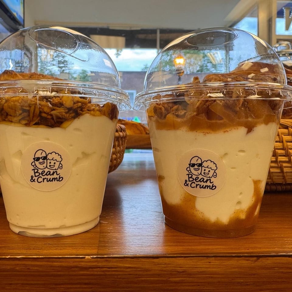 Two Bean & Crumb dessert cups layered with creamy filling and crunchy toppings sit on a wooden counter.