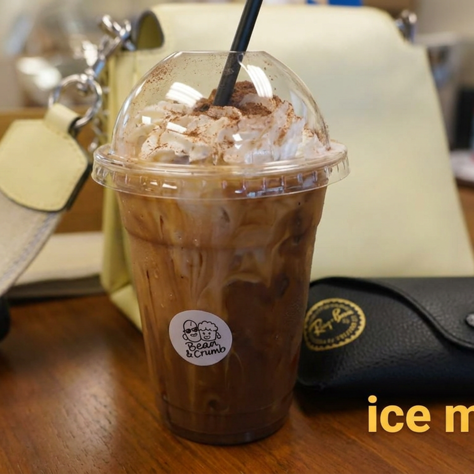 Iced mocha topped with whipped cream and cocoa sits on a wooden café table beside a hat and black wallet.